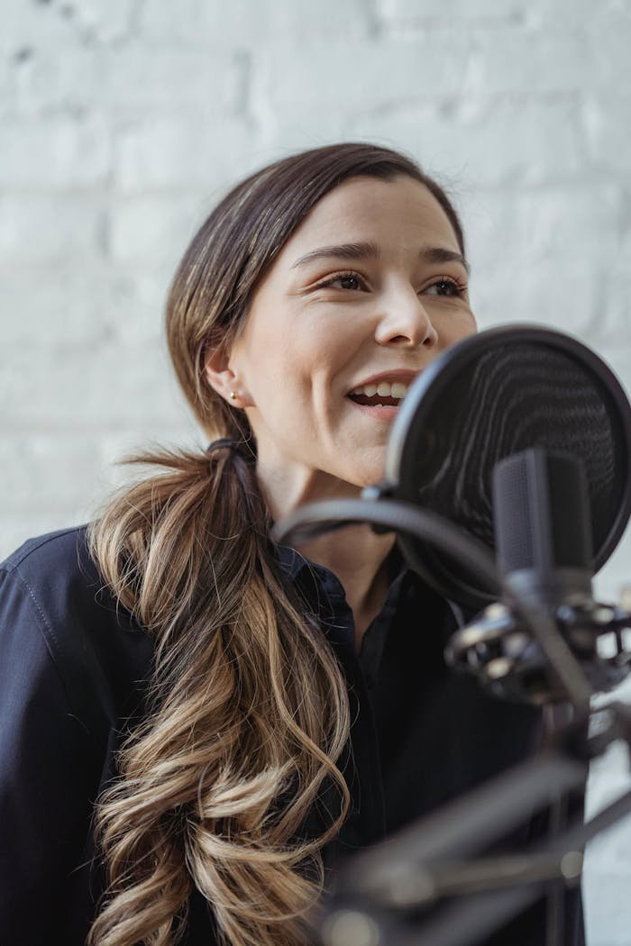 menu-08 Smiling woman in casual attire speaking into a microphone in a modern studio setting.